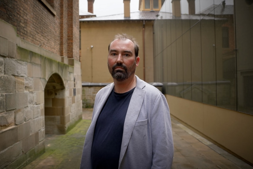 A man with a blue shirt and grey jacket stands among sandstone buildings