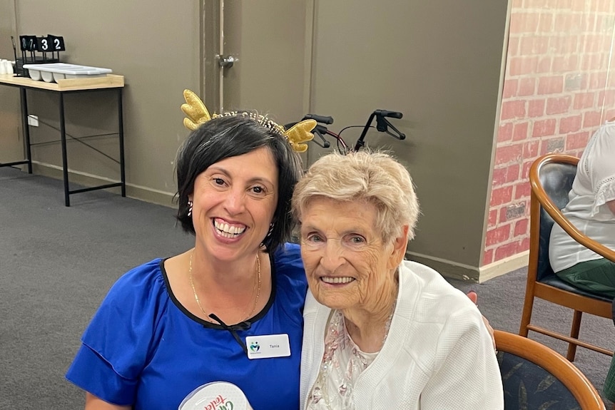 A middle aged woman with dark hair and a reindeer antler headbands smiles at the camera. She's sitting next to an elderly woman.