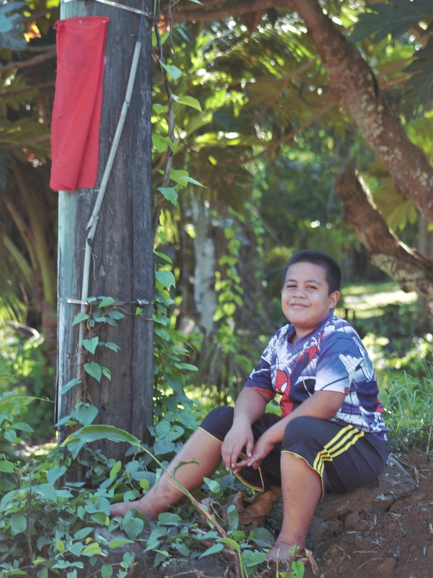Young boy sits on rocks next to a telephone pole with red fabric tied around it.