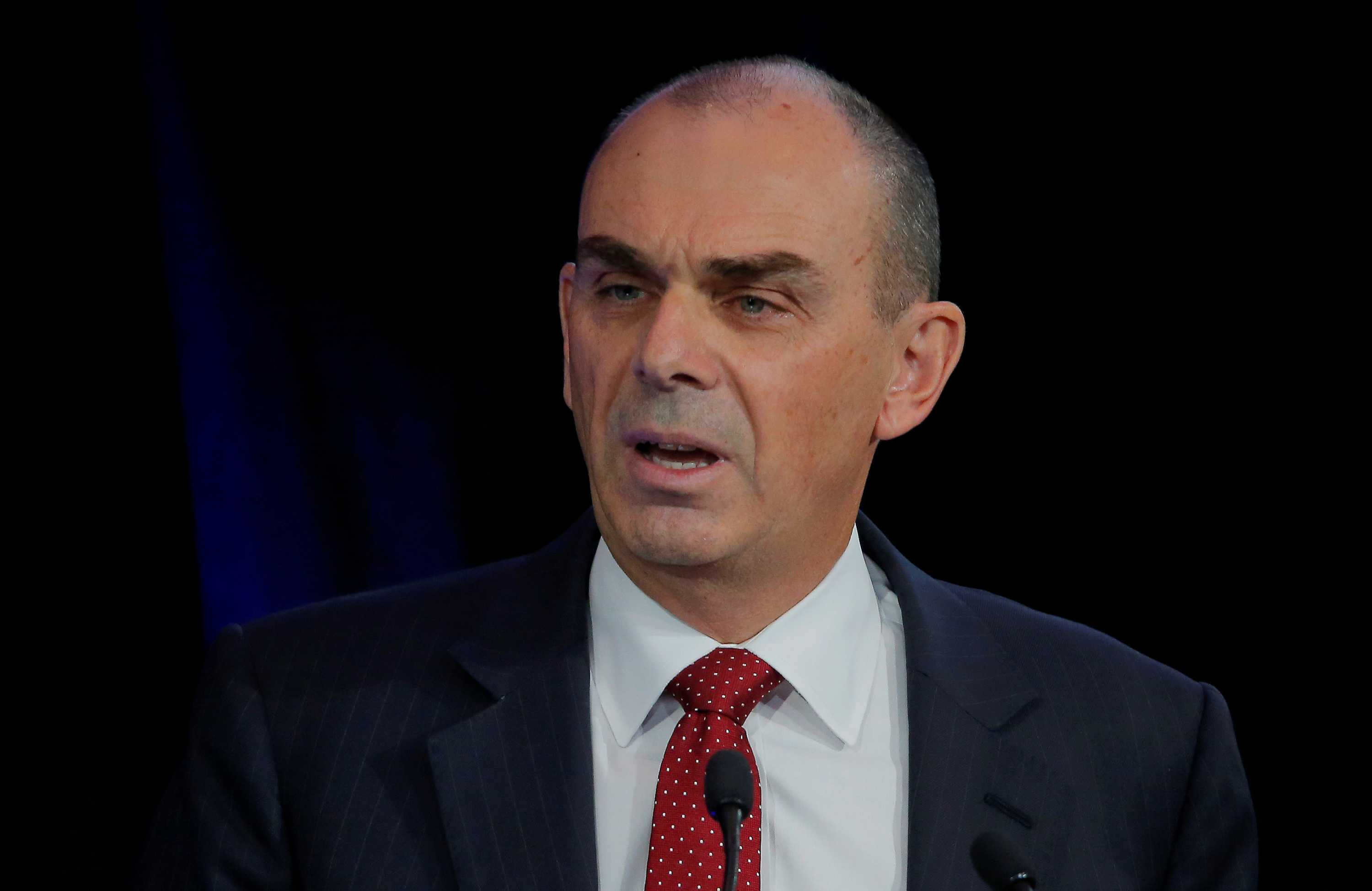 A head-and-shoulders shot shows a middle-aged white man in a dark suit and red tie with short cropped dark hair