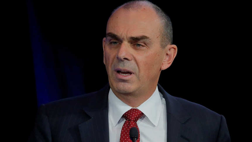 A head-and-shoulders shot shows a middle-aged white man in a dark suit and red tie with short cropped dark hair