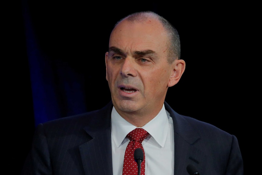 A head-and-shoulders shot shows a middle-aged white man in a dark suit and red tie with short cropped dark hair