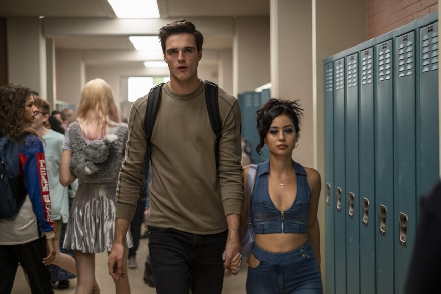 A tall teenage boy walks through the school hallway hand in hand with his shorter girlfriend in a revealing outfit.