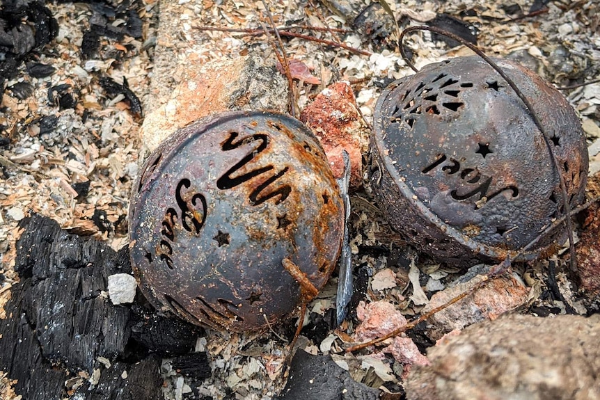 Two burnt metal Christmas baubles with the words 'Noel' and 'Peace' lying in rubble