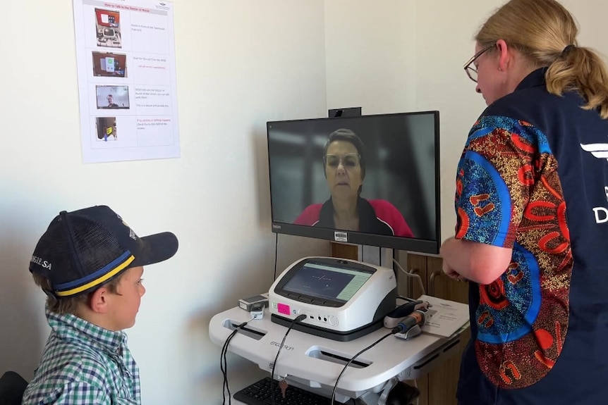 A young boy wearing a hat sits in front of a computer screen showing a livestream of a doctor.