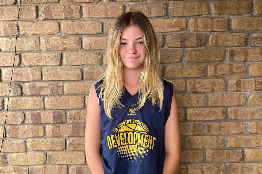 A teenage girl stands in front of a brick wall wearing her SA country basketball development kit.