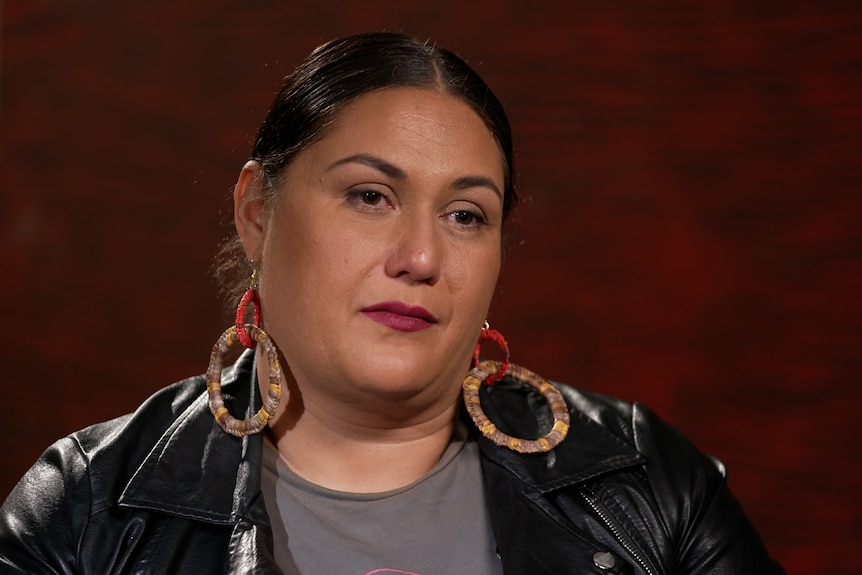 A woman in a black leather jacket with black hair, big earings and red background
