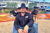 Murray Wilkinson sits between his dogs. The dogs have their tongue out looking at the camera. 