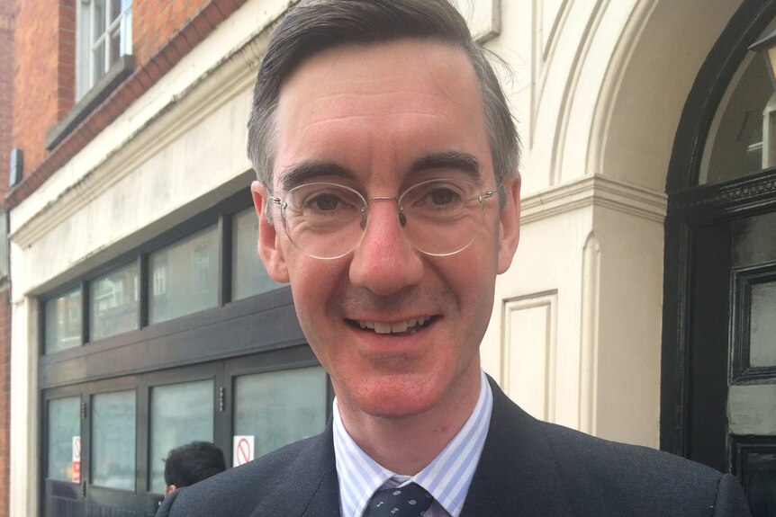 Jacob Rees-Mogg poses for a photo in the street, holding files and wearing a suit.