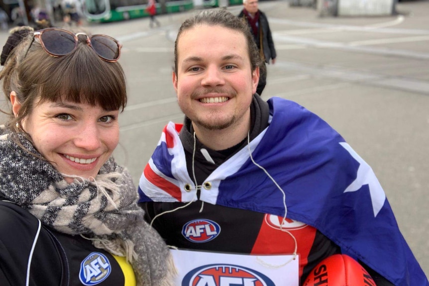 A woman stands next to a man wearing an Australian flag around his shoulders and holding and AFL ball.