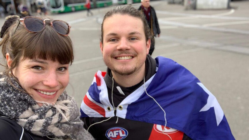 A woman stands next to a man wearing an Australian flag around his shoulders and holding and AFL ball.
