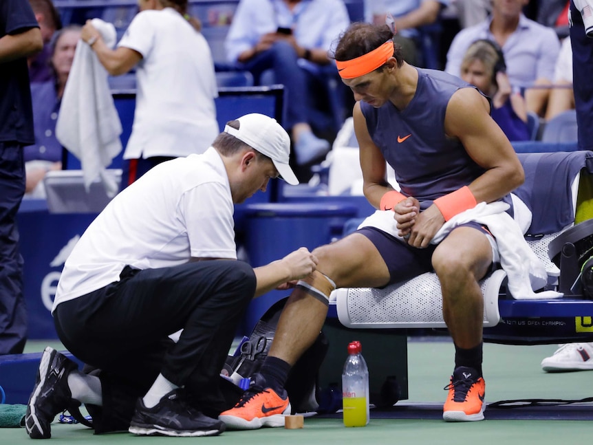 A male tennis player sits on a chair and has his knee strapped by a man in a white shirt
