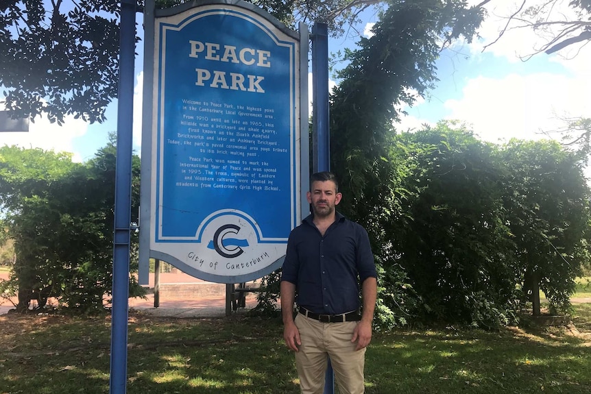 A man in a blue shirt and beige chinos standing near a blue sign.