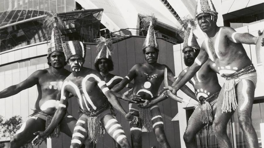 Group of Aboriginal men wearing traditional body paint and costume pose infront of Sydney Opera House