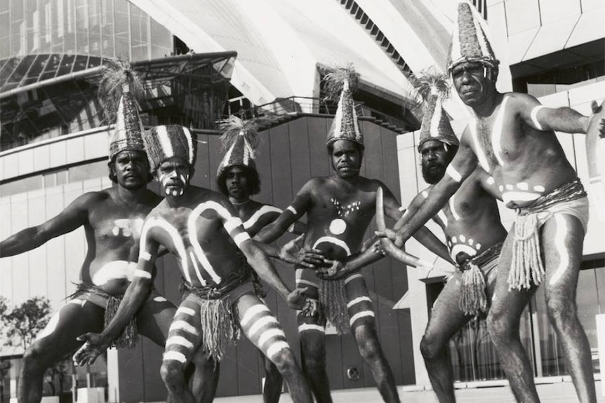 Group of Aboriginal men wearing traditional body paint and costume pose infront of Sydney Opera House