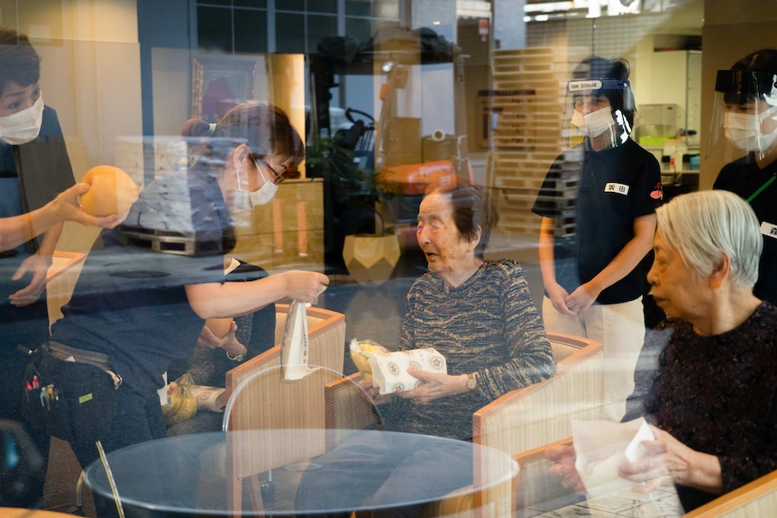 Two women sit in a chair as two masked women offer them food.