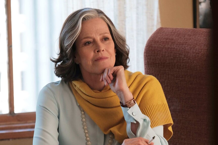 Sigourney Weave sits at office desk dressed stylishly, with yellow wool sweater around her shoulders, resting chin on one hand.