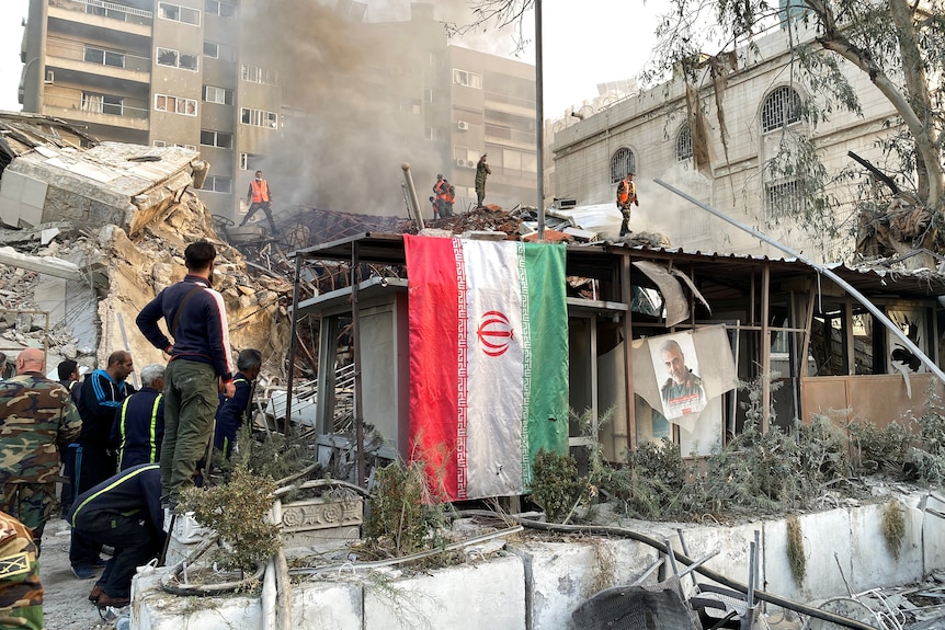 Smoke rises from a building surrounded by rubble as people look on