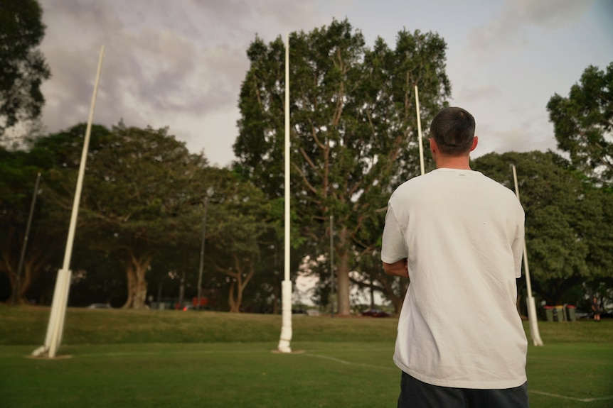 A young man in a white sirt looking at some AFL goal posts