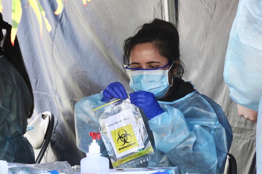 A woman sits at a table under a gazebo wearing PPE gear and closing a specimen bag.