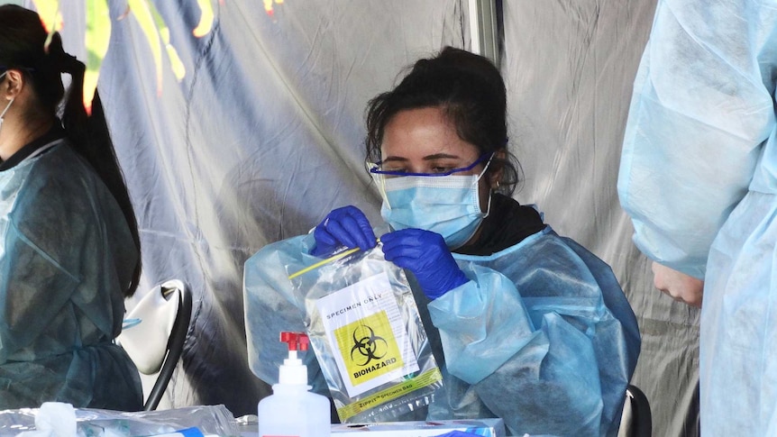A woman sits at a table under a gazebo wearing PPE gear and closing a specimen bag.
