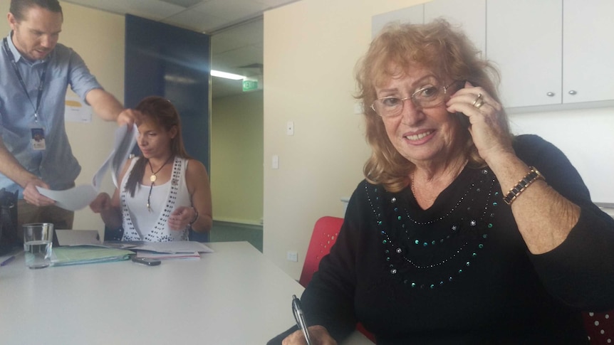 An older volunteer holds mobile phone to ear and smiles at camera. Officials working in the background.