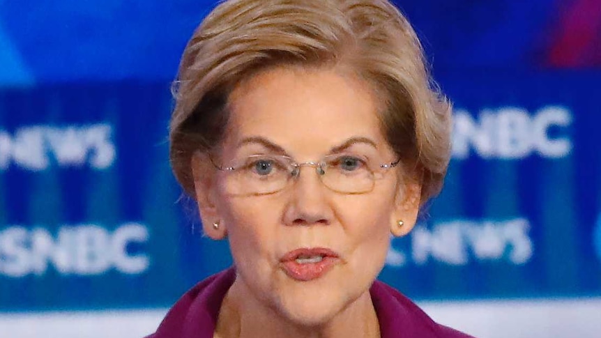 Elizabeth Warren, wearing a purple blazer, stands at a lectern talking