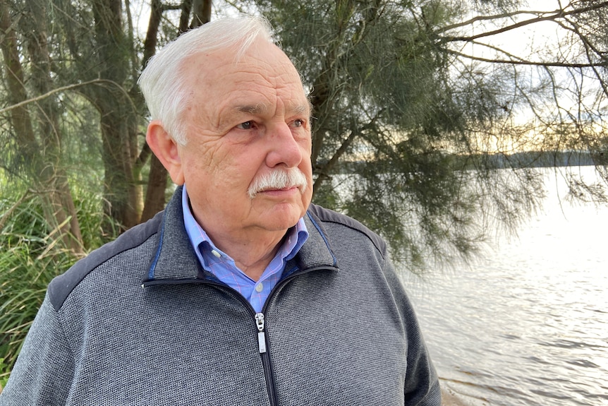 A white-haired man stands near a lake.