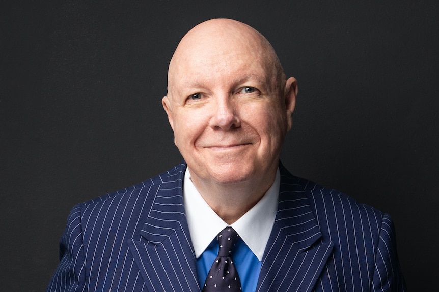 A bald middle-aged man wearing a blue pinstripe suit and polka dot dark tie smiles at camera.
