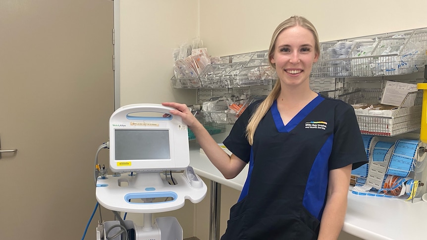 A woman in a nurses uniform and long blonde hair smiles at the camera, medical equipment around her