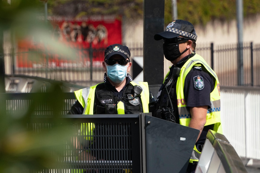 Trees in the foreground, two Qld police officers in masks in the background.