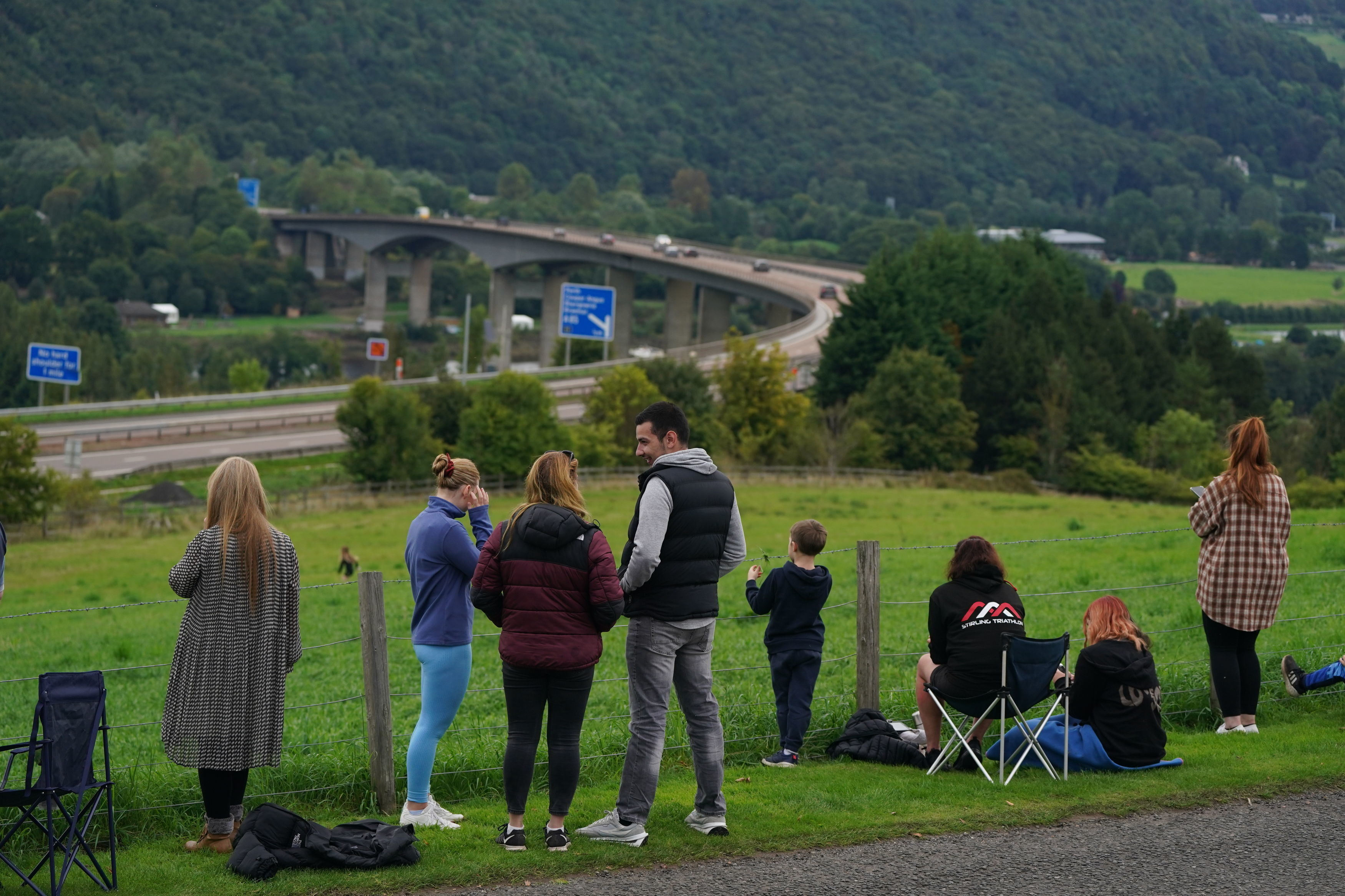 Crowds stand on a grassy hillside looking out at a highway. 