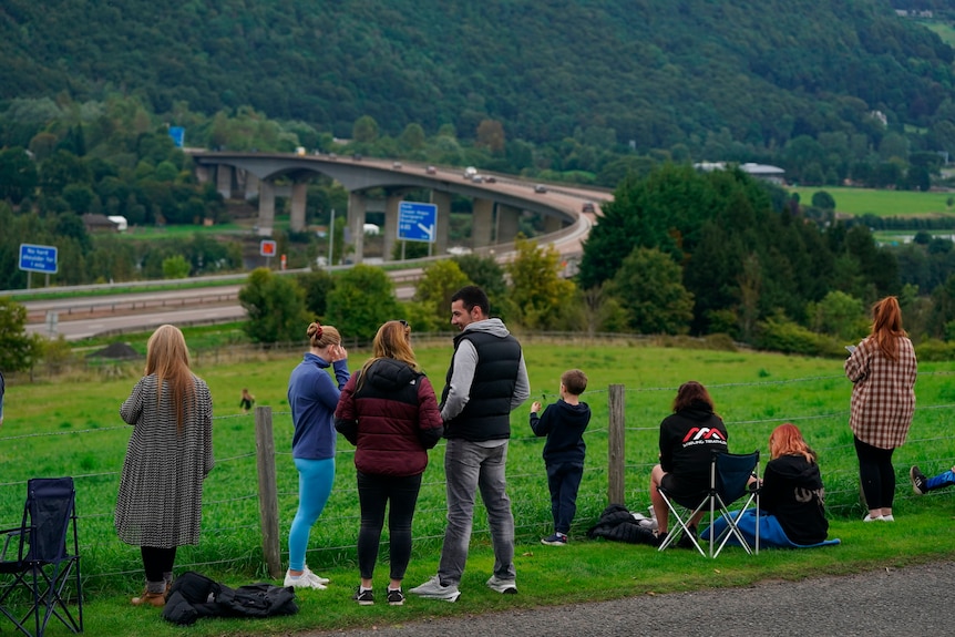 Crowds stand on a grassy hillside looking out at a highway. 