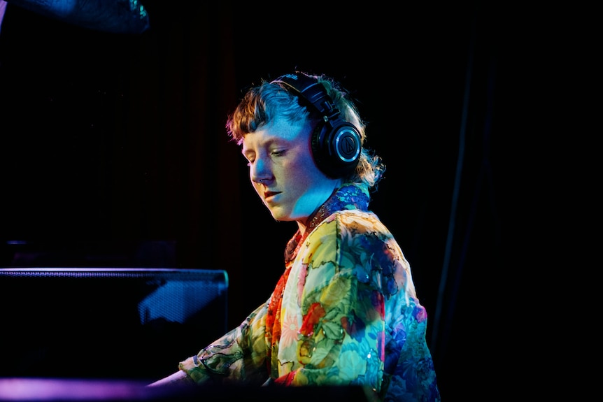A non-binary person DJs in a dark nightclub with some coloured lighting. 