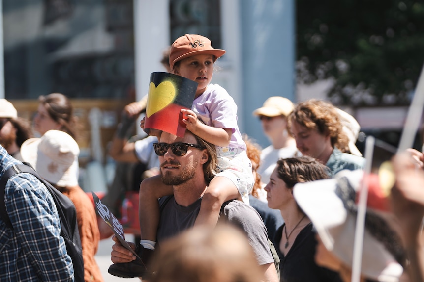 A child sitting on a man's shoulder holding an Aboriginal flag where the yellow circle in the centre is shapes as a heart.