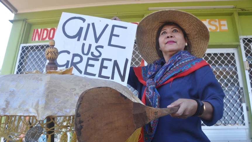 closeup image of woman holding wooden spoon and sign reading: "Give us green" outside her restaurant.