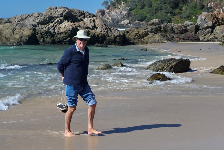 An older man wearing jeans, shirt and a hat, stands on a beach smiling, with rocks in the background.