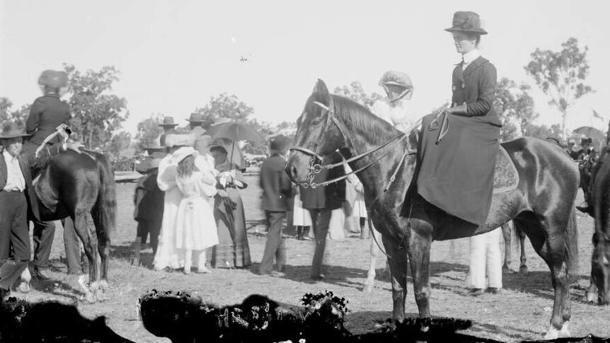 Black and white image of a woman sitting side saddle on a horse wearing a very formal dress and hat