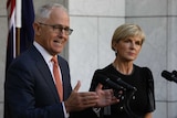 Malcolm Turnbull gestures with both hands towards the microphone. Julie Bishop stands beside him, watching the crowd.