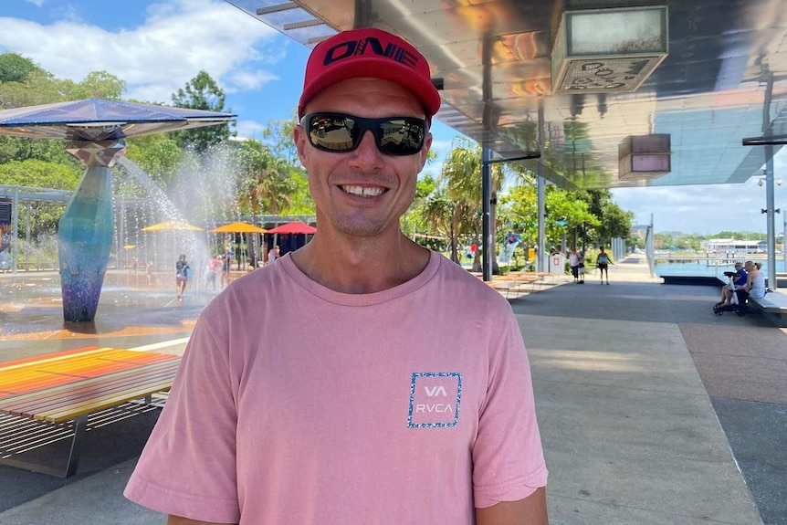 A man wearing a hat and sunnies in front of a water play area smiles at the camera.