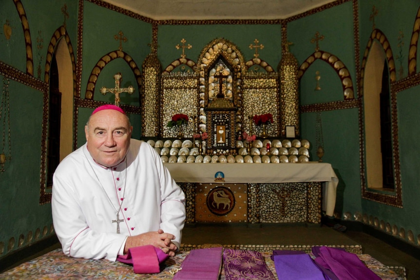 A large man wearing a skullcap and white robe in front of a pearl shell altar in a church.