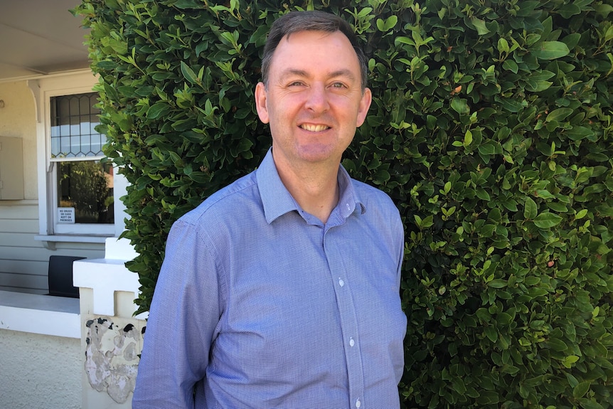 A man stands in front of bushes smiling at the camera.