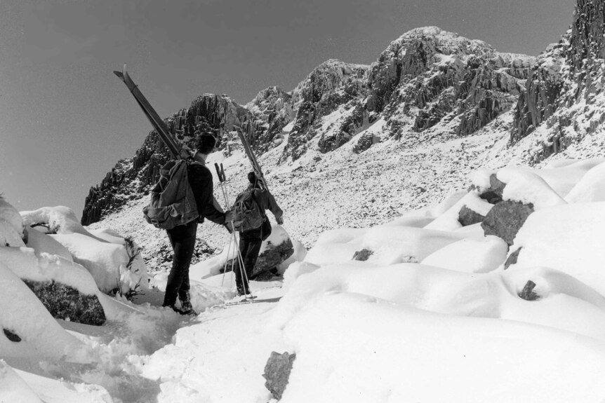 Black and white photo of two skiiers with skis on shoulder walking to summit of snowy mountain