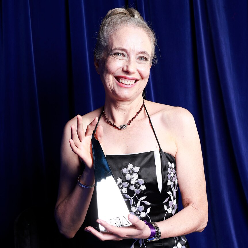 Sue Lowry holds a pyramid-shaped ARIA award and smiles at the camera.She wears a black formal dress with white flower embroidery