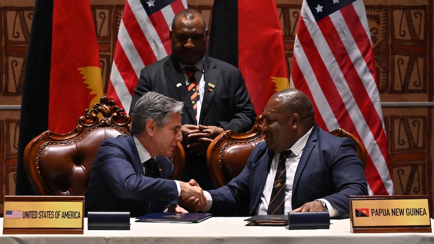 Win Bakri Daki and Antony Blinken shake hands sitting at a dark timber desk with a man standing behind near US and PNG flags.