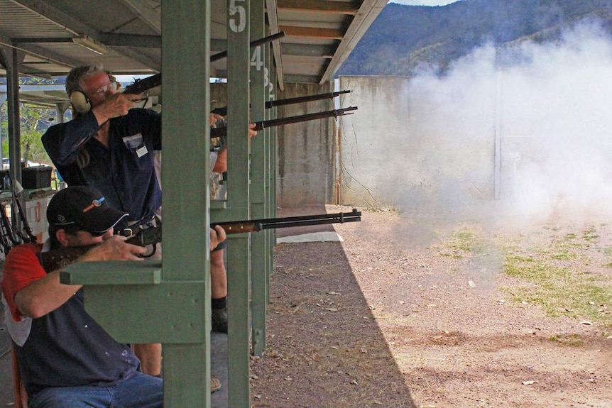 Competitors lining up side by side and shoot their rifles. A puff of white smoke comes out the barrels of their guns.