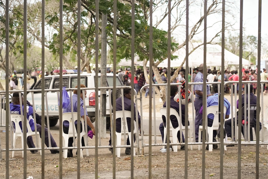 A group of PNG police sit on chairs near a fence as they watch over a crowd of people.