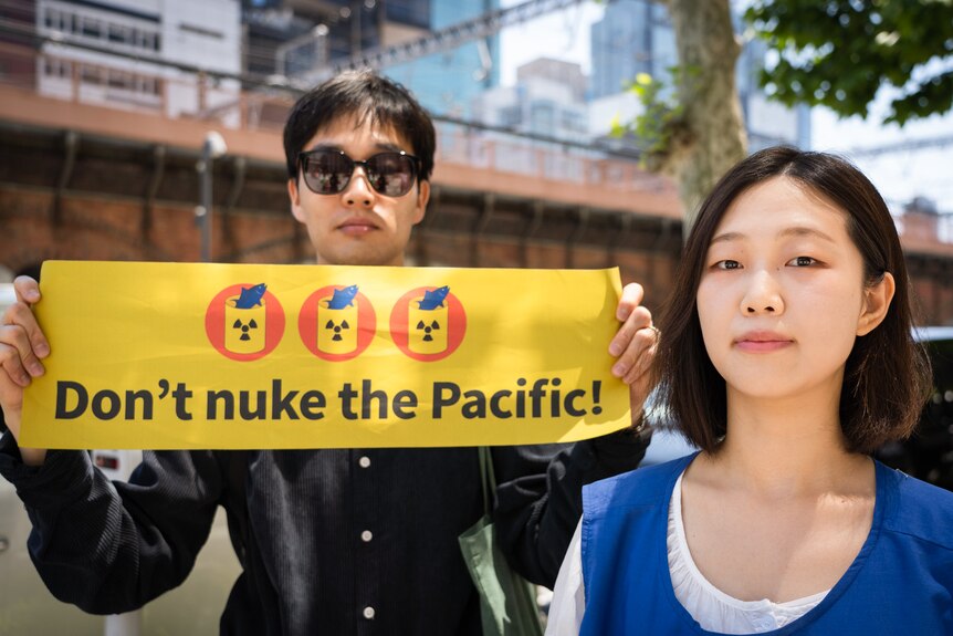 A man wearing sunglasses holds a sign saying Don't nuke the Pacific while a woman stands beside him.
