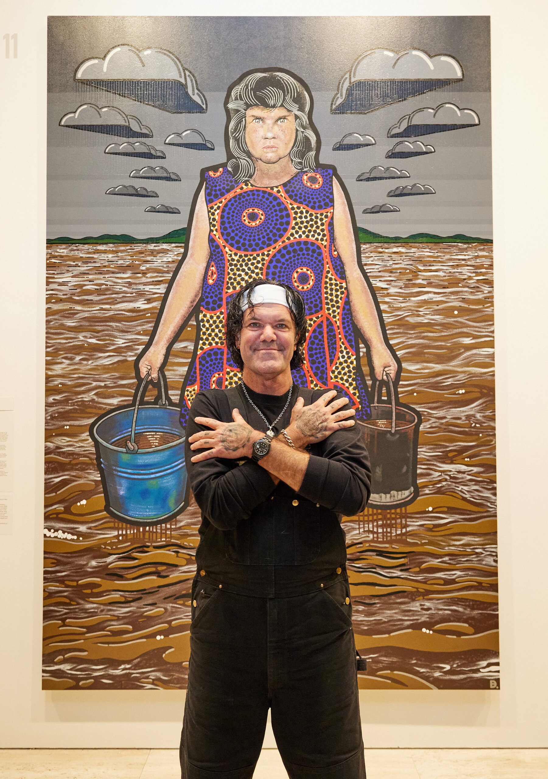 An Indigenous man smiles, arms crossed over his chest, while standing in front of a large portrait of an Indigenous woman