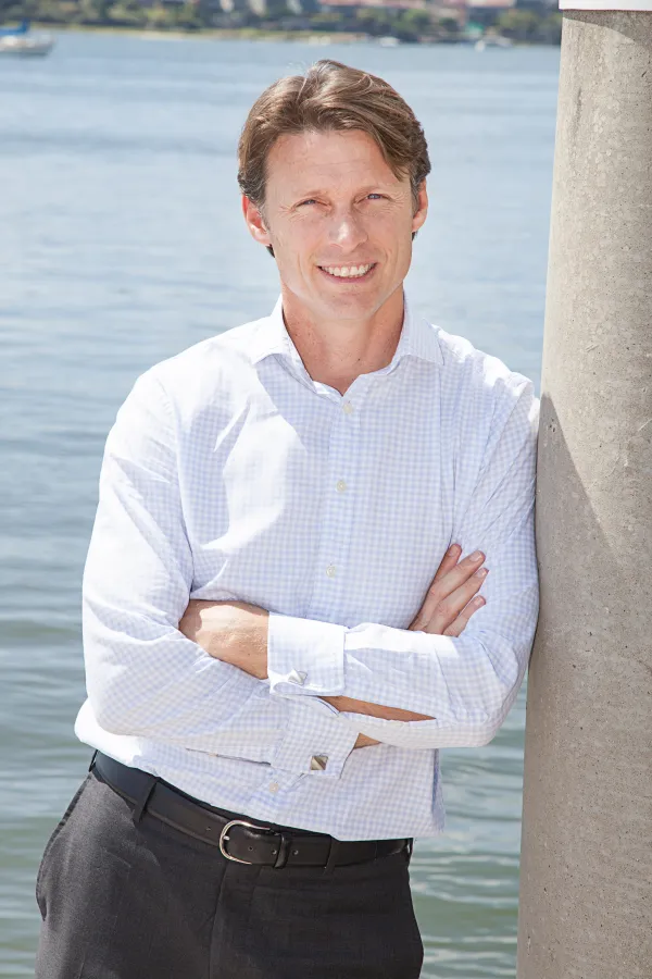 A smiling blonde man in a business shirt leans against a pole in front of a body of water with his arms crossed.